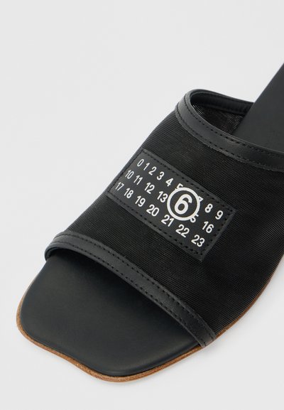 Μαύρο σανδάλι με φαρδιά λουρί από δίχτυ, που διαθέτει ραμμένη ετικέτα με αριθμούς και έναν κύκλο με τον αριθμό 6.