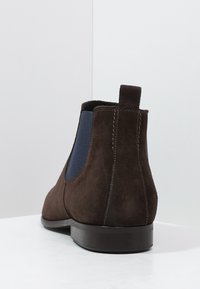 Bottes Chelsea en daim marron avec panneaux élastiques latéraux bleus, talon bas empilé, et une languette à l'arrière. Des coutures nettes accentuent le design.