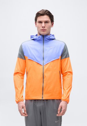 Mann trägt eine Windjacke mit Farbeinsätzen in Blau, Orange und Grau, steht mit entspannten Händen seitlich an einem einfachen Hintergrund.