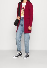 Blazer long bordeaux sur un t-shirt graphique blanc, associé à un jean taille haute bleu clair et des baskets noires à semelle basse. Sac à main noir.