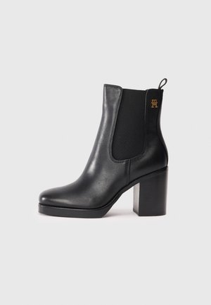 Bottine en cuir noir avec talon carré, panneaux latéraux élastiques, languette de tirage, et petit logo doré près du sommet sur fond gris clair uni.