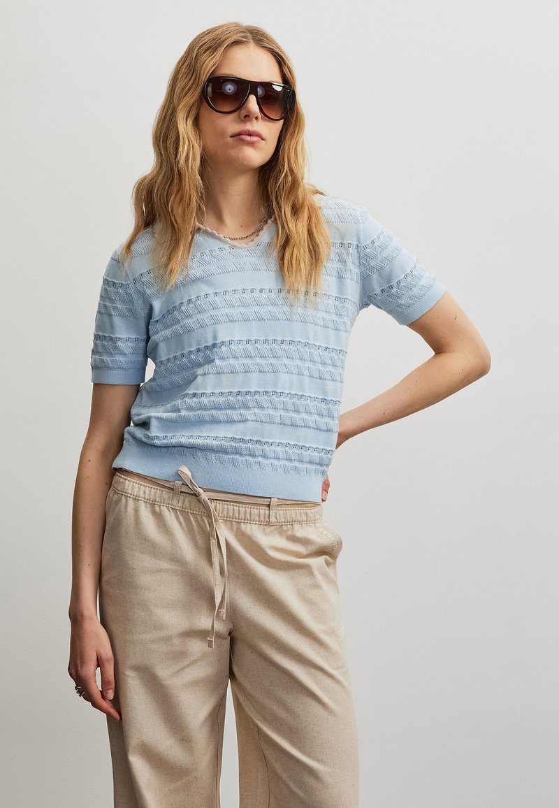Femme aux longs cheveux ondulés portant des lunettes de soleil foncées, un haut tricoté bleu clair à manches courtes et un pantalon beige avec cordon de serrage, debout avec une main sur la hanche.