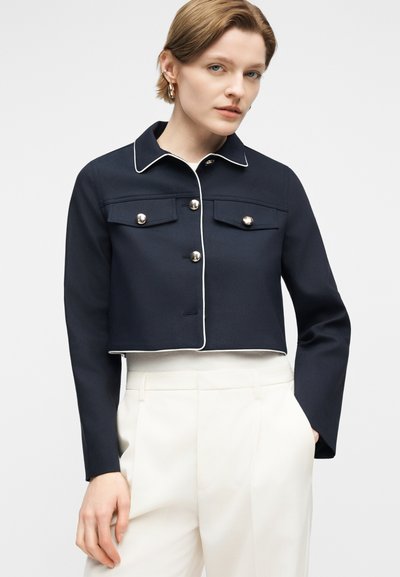 Γυναίκα που φοράει cropped μπλε σκούρο σακάκι με ασημένια κουμπιά και λευκή λεπτομέρεια ρέλι, σε συνδυασμό με λευκό παντελόνι ψηλόμεσο, ποζάροντας σε απλό φόντο.