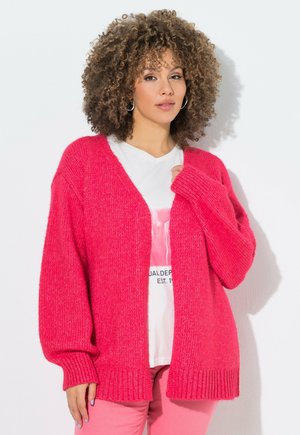 Kobieta o kręconych włosach, nosząca kolczyki koła, jasnoróżowy kardigan, białą bluzkę z nadrukiem oraz jasnoróżowe spodnie na jednolitym białym tle.