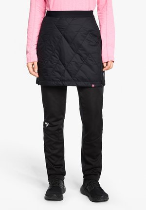 Person trägt einen gesteppte schwarzen Rock über schwarzen Hosen, ein rosa Langarmoberteil und schwarze Sportschuhe, steht vor weißem Hintergrund.