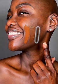 Modell med en jämn, mörk hy visar tre makeup-prover – två i rika bruna nyanser och en genomskinlig. Hon pekar med en välvårdad hand.