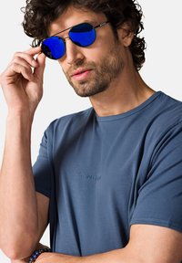 Niebieska koszula z krótkim rękawem z subtelnym logo, o gładkiej fakturze. Osoba nosi lustrzane niebieskie okulary przeciwsłoneczne, co podkreśla sportowy wygląd.