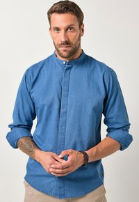 Man met een baard en een tatoeage, gekleed in een blauwe mandarin colbert met opgerolde mouwen, met zijn handen voor zich samengevoegd voor een effen lichte achtergrond.