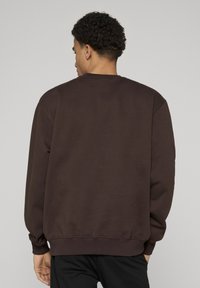 Sweatshirt marron à col rond, manches longues et poignets côtelés. Texture douce et coupe décontractée, vue uniquement de dos.