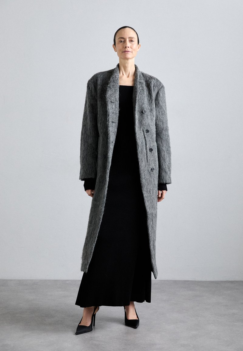 Long manteau en feutre gris avec de grands boutons, porté sur une robe noire à côtes, accompagné de chaussures à talons hauts noires. Surface lisse, design structuré.