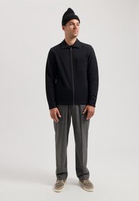 Chaqueta negra con cremallera, cuello y bolsillo frontal, combinada con pantalones grises con pliegues y zapatillas beige, llevada con un gorro negro.