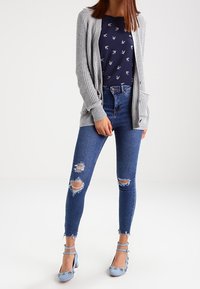 Šedý pletený kardigan přes námořnicky modrou košili s bílým potiskem, doplněný o opotřebované modré džíny a světle modré lodičky s květinovými detaily.