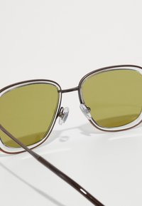 Brūnas metāla saulesbrilles ar caurspīdīgām akcentiem, zaļiem lēciem un deguna spilventiņiem. Liels, leņķisks rāmja forma ar elegantu dizainu un plānām kājām.