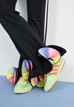 Person in schwarzen Hosen mit regnbuntem Rüschensaum und hellgrünen Sneakers mit orangefarbenen Streifen, steht auf einem blauen Teppich.