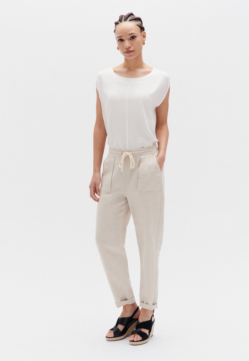 Crème blouse met zachte draping, gecombineerd met lichtbeige op maat gemaakte broeken met zakken en een trekkoord in de taille. Zwarte wiggen sandaaltjes maken de look af.
