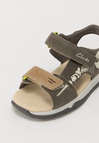 Grå och brun sandal med kardborreband, vadderad fotbädd och mönstrad design. Har en gummisula och accentfärger i grönt.