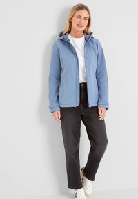Light blue softshell jacket with a hood, zip closure, and side pockets, paired with black jeans and white sneakers. Smooth texture.