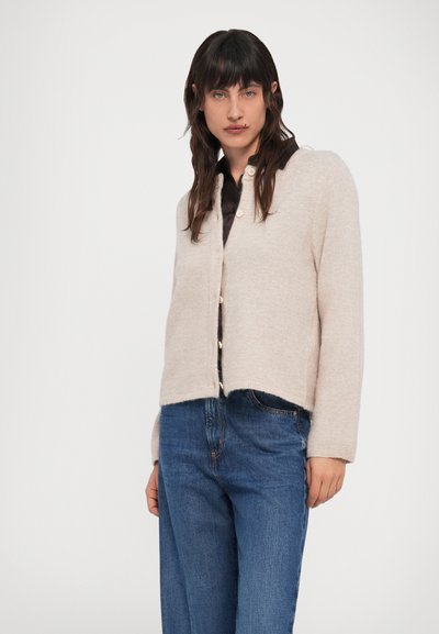 Femeie cu păr lung și închis la culoare, purtând un cardigan bej cu nasturi peste o cămașă neagră și blugi albaștri, stând în fața unui fundal alb simplu.