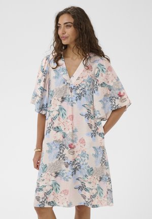 Kobieta z kręconymi włosami ubrana w sukienkę o długości do kolan z dekoltem w serek, szerokimi rękawami i pastelowym kwiatowym wzorem, pozująca z jedną ręką w kieszeni.