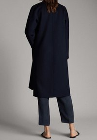 Lungo cappotto blu navy dalla linea dritta, con maniche lunghe e collo alto, abbinato a pantaloni scuri e sandali piatti. Finitura in tessuto liscia.