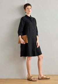 Robe en lin noir à boutons, avec des manches trois-quarts, une poche avant et des fentes sur les côtés. Assortie à des sandales brunes texturées et à une pochette beige.