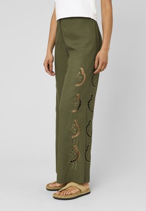 Femme portant un pantalon large vert olive avec des découpes en forme de croissant de lune, des sandales à plateforme beiges et un haut blanc sans manches.