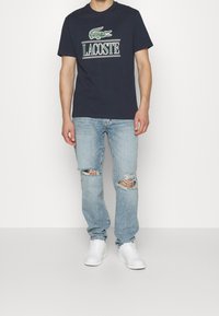 Man wearing navy Lacoste t-shirt, light blue ripped jeans, and white sneakers standing against a plain white background.