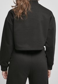 Person som bär en svart kortärmad sweatshirt och matchande svarta byxor, visad bakifrån med löst hår över ena axeln.