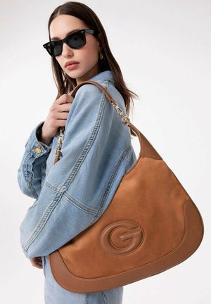 Donna che indossa occhiali da sole neri e una giacca di jeans azzurro chiaro, porta una grande borsa a tracolla in pelle marrone con un emblema "G".