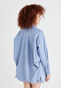 Cămașă albastră cu dungi roșii și albe, având o croială lejeră, mâneci lungi și un material moale, texturat. Funda de cămașă și nasturi vizibili.