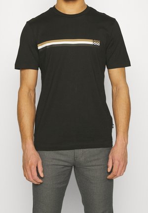 Man draagt een zwart T-shirt met korte mouwen met horizontale beige, witte en grijze strepen en de tekst "BOSS" op de borst, gecombineerd met een grijze broek.