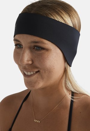 Young woman with light skin and blonde hair wearing a black headband and a thin gold necklace, smiling slightly, against a white background.