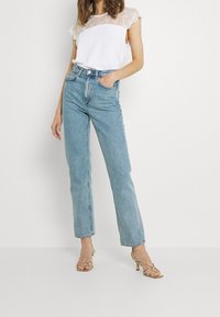 Leichtes weißes Oberteil mit Spitzen-Schulterdesign, kombiniert mit hoch taillierten, geraden hellblauen Jeans und goldenen Riemchen-Heels.