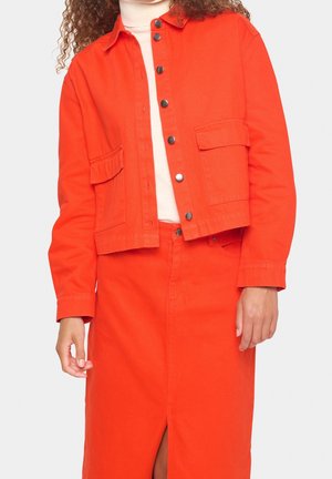 Mujer vistiendo una chaqueta corta abotonada de color naranja brillante y una falda de cintura alta a juego con una abertura frontal, sobre un suéter de cuello vuelto color crema.