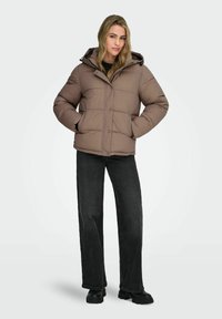 Veste d'hiver matelassée marron avec capuche, présentant un design matelassé, des boutons à l'avant et des poches latérales, associée à un jean noir et des bottes noires.