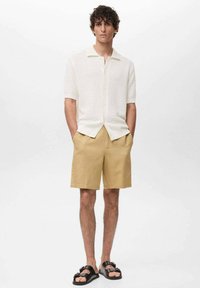 Kortärmad vit texturerad skjorta med krage och knäppning, matchad med beige shorts och svarta sandaler.
