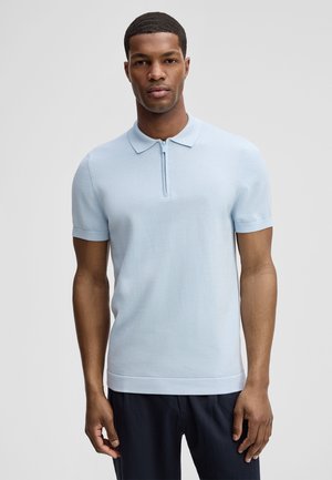 Mężczyzna ubrany w jasnoniebieską, krótko rękawową koszulkę polo z przednim zamkiem błyskawicznym, stojący na tle jednolitego szarego tła.