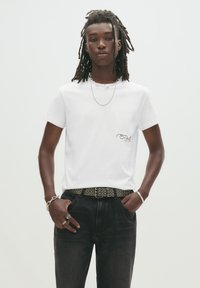 Hvid bomulds t-shirt med korte ærmer, der har et lille logo på brystet, parret med mørke denim jeans og et dekorativt bælte.