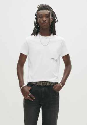 Hvid bomulds t-shirt med korte ærmer, der har et lille logo på brystet, parret med mørke denim jeans og et dekorativt bælte.