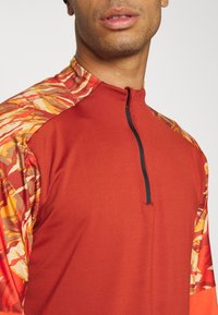 Rotes Langarmhemd mit einem schwarzen Reißverschluss. Die Ärmel zeigen ein lebhaftes orange-gelbes Camouflage-Muster. Hergestellt aus glattem, dehnbarem Stoff.