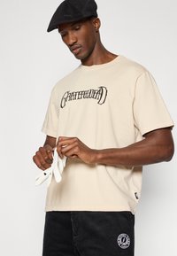 Μπεζ βαμβακερό μπλουζάκι με μαύρο κείμενο "Grateful Dead", συνδυασμένο με μαύρο παντελόνι κορντό υφής με στρογγυλή λογότυπη κουμπότρυπα.