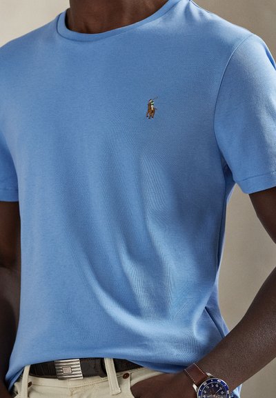 Šviesiai mėlynas medvilninis marškinėlis su apvalia iškirpte ir trumpomis rankovėmis. Kairėje krūtinės pusėje siuvinėtas logotipas. Dėvimas su rudos spalvos diržu ir laikrodžiu.