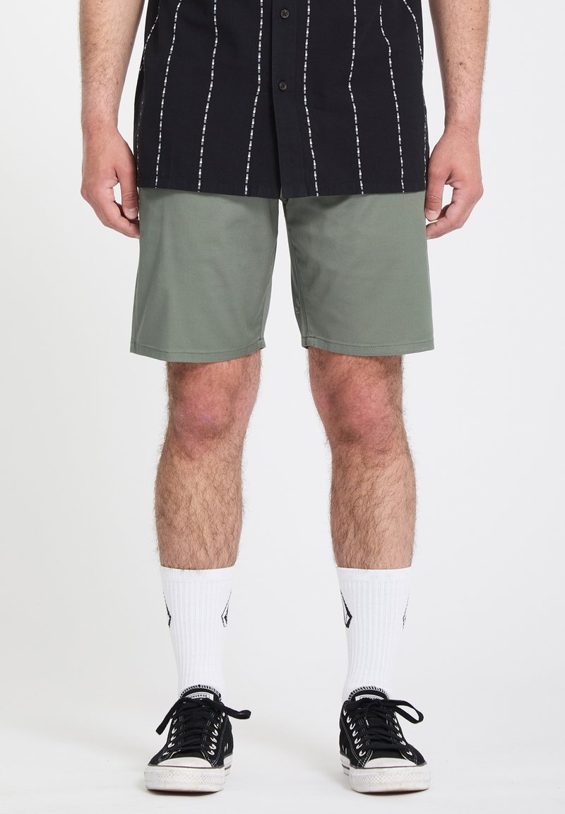 Homme portant un short vert olive, des baskets noir et blanc, des chaussettes blanches, et une chemise noire à manches courtes avec des rayures blanches verticales.