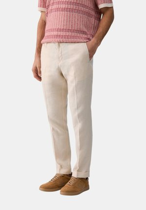 Uomo che indossa pantaloni beige e sneakers marroni con le mani nelle tasche, mostrato dalle spalle ai piedi su uno sfondo semplice.