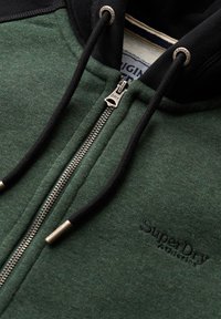 Sudadera verde con cremallera y capucha negra, que cuenta con una cremallera de metal, cordones y un logo sutil en la parte delantera. La textura es suave y casual.