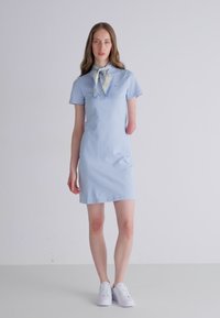 Lys blå polodress med korte ærmer, med krave og et mønstrete skjerf. Kombinert med hvite sneakers, enkel og tettsittende design.