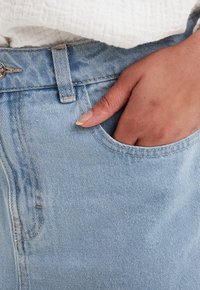 Jeans de mezclilla azul claro con una textura suave, bolsillo visible y cierre de botón metálico en la cintura. Mano descansando en el bolsillo.