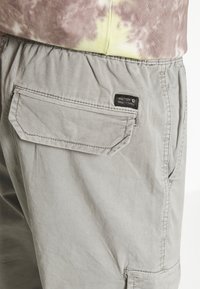 Pantaloni corti grigio chiaro con una tasca a soffietto sul lato e una piccola etichetta nera con la scritta "NINETEEN NINETY THREE" sotto l'orlo di una maglietta tie-dye.