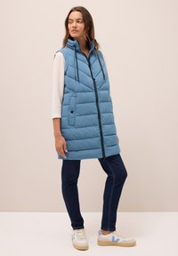 Femme portant un gilet sans manches matelassé bleu, un haut à manches longues blanc, un jean foncé et des baskets blanches avec des accents bleus, se tenant devant un fond beige.