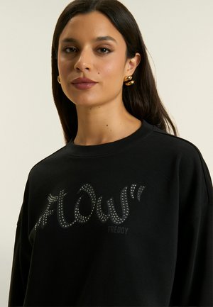 Donna con lunghi capelli scuri indossa una felpa nera con la scritta "FLOW" borchiata e orecchini a cerchio dorati su sfondo beige.